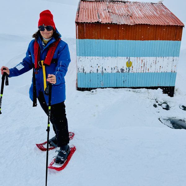 La autora con raquetas de nieve junto a caseta con bandera argentina en Antártida (límite IAATO 100).